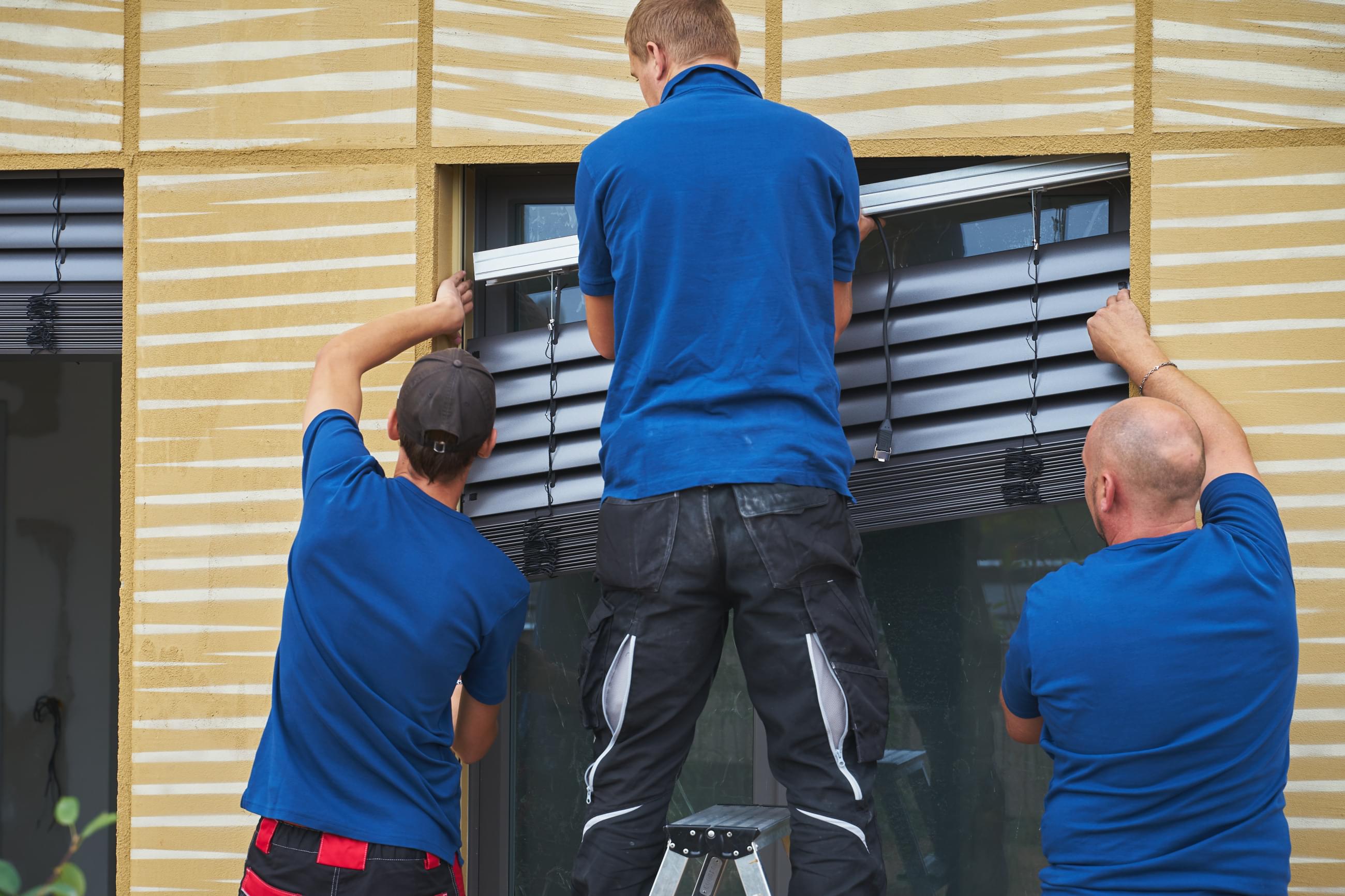Tři montážníci zasazují žaluzie do prostoru okna